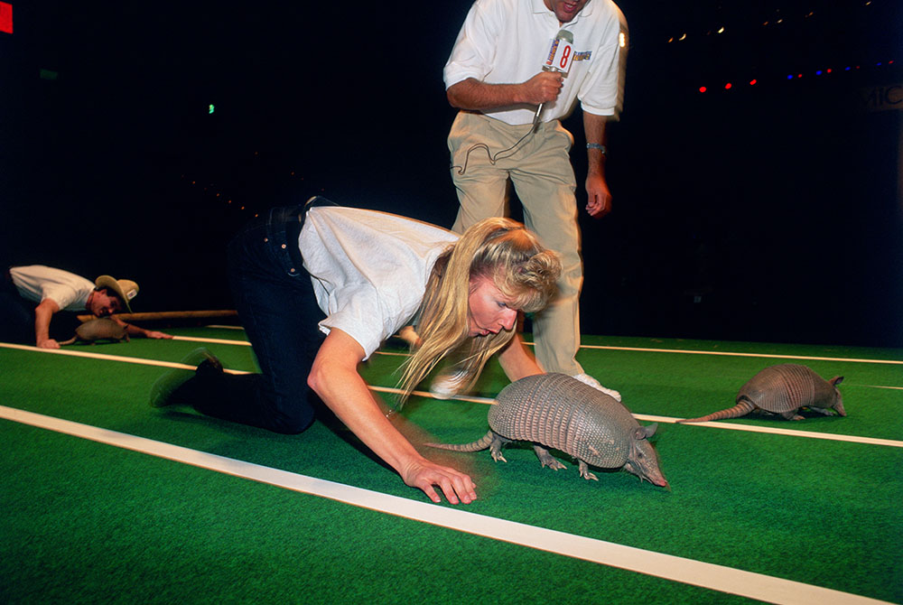 A blonde woman in jeans and a white t-shirt crawls on a green field, mirroring a nearby armadillo, while another person and armadillo are visible in the background