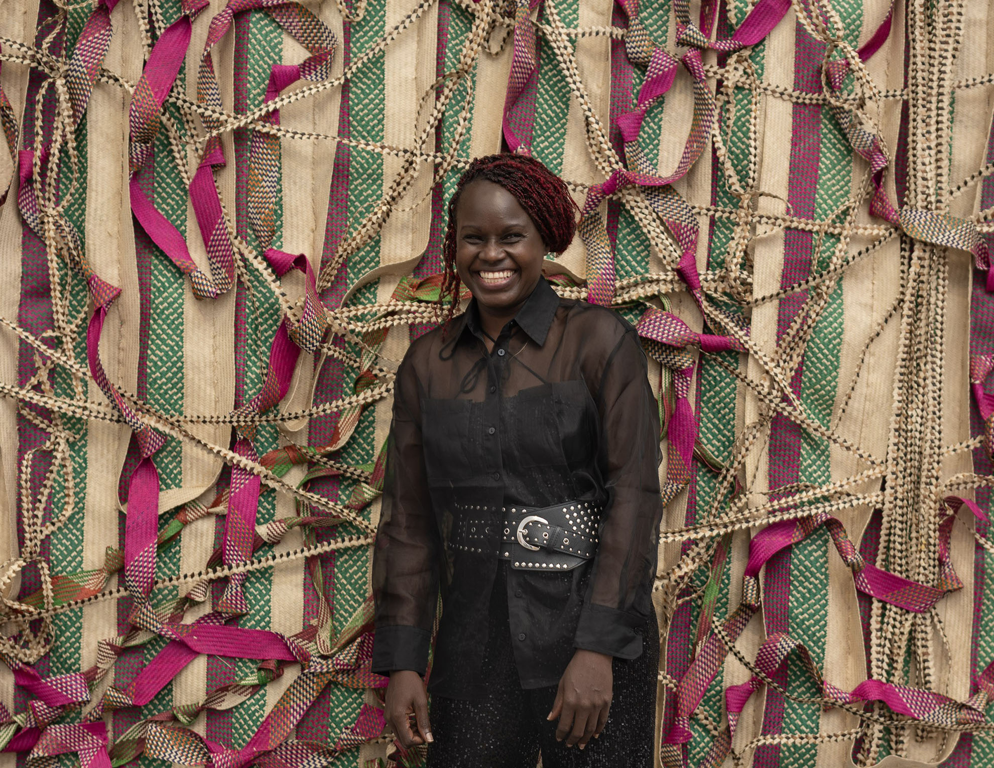 Portrait of artist Acaye Kerunen standing in front of a large woven wall tapestry made of natural fibers and bright pink and green stripes.