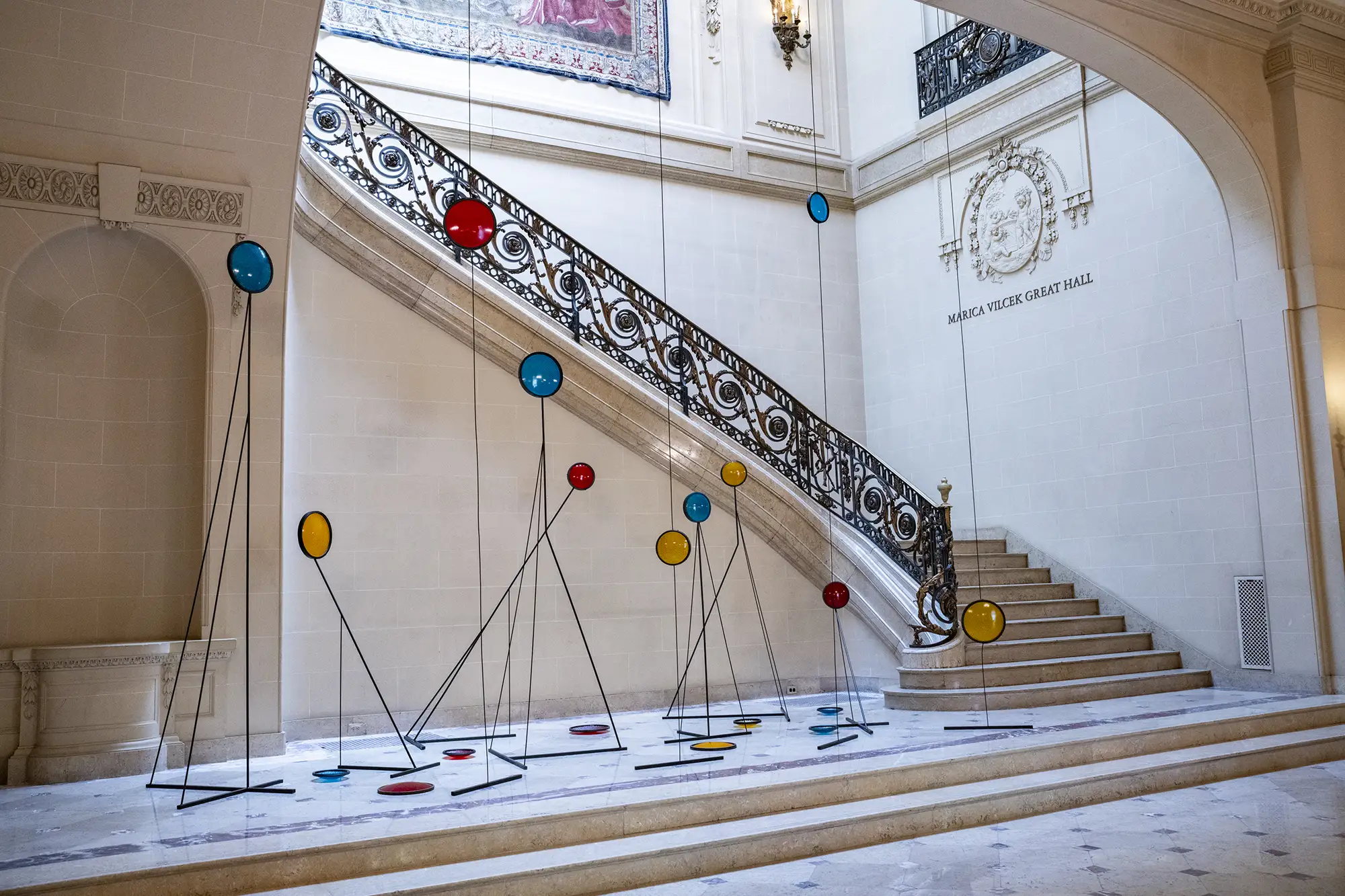 A modern art sculpture featuring colorful, glass-like discs hangs from the ceiling and stands on the floor in a grand, ornate marble entryway with a large staircase. The discs are in various sizes and colors, including red, yellow, and blue. They are suspended by thin, black wires that also hold up some of the discs from the floor. The staircase has a decorative, black wrought-iron railing with gold accents.