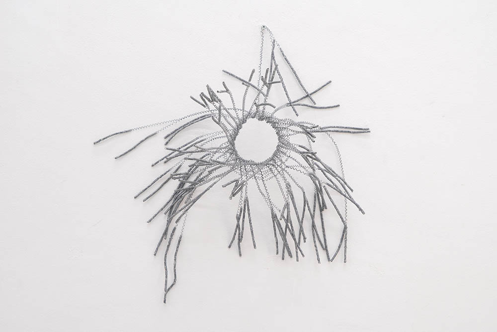 Wall-mounted woven wire sculpture featuring a central ring with grey wire tendrils branching outward against a plain white wall.