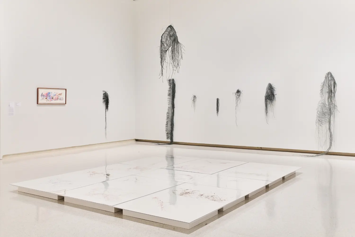 Art gallery featuring a low tiled floor platform with subtle drawings and delicate wire sculptures hanging along white walls.