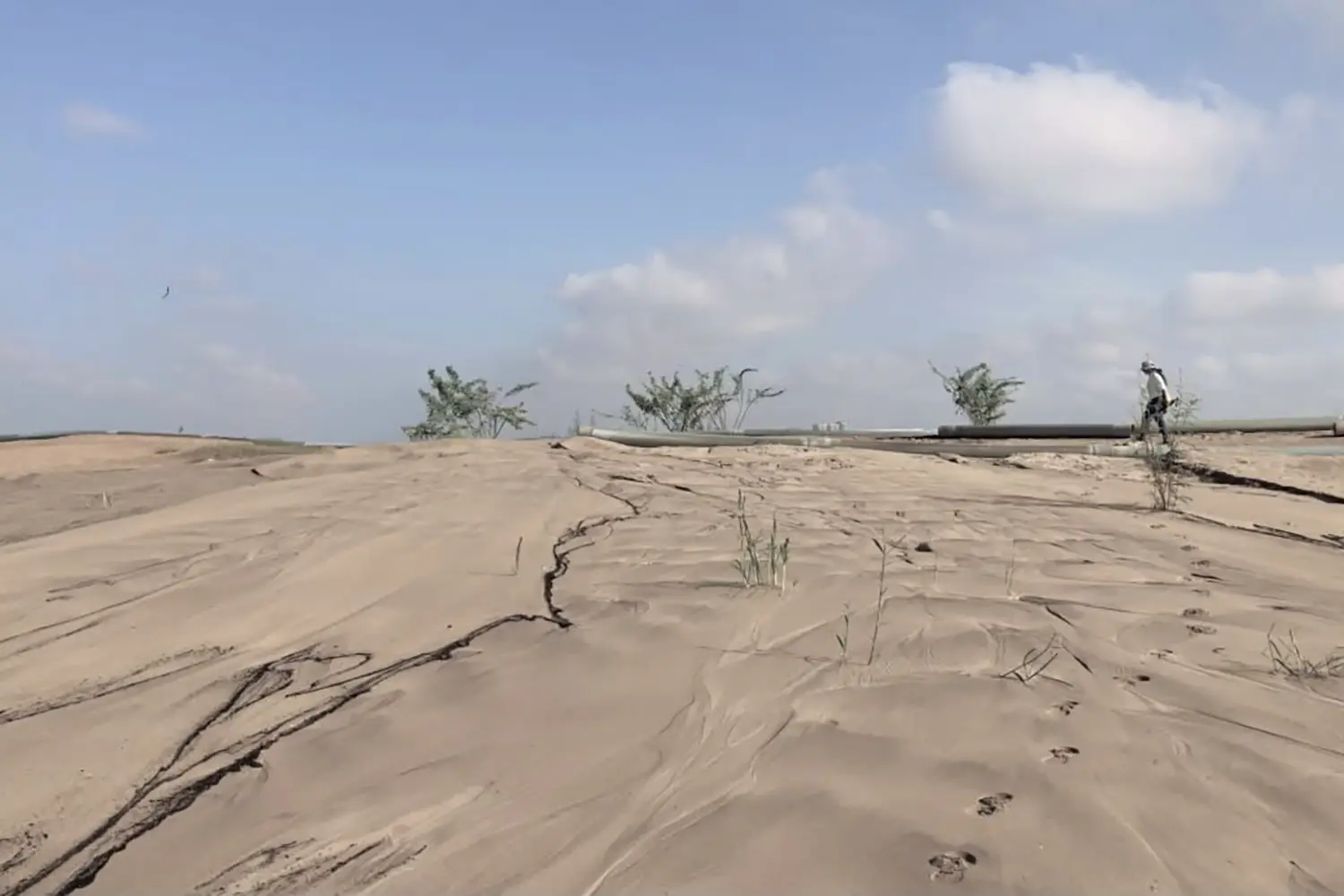 Desolate sandy ground with dark cracked earth, sparse vegetation, industrial pipes, and a person walking in the far distance.