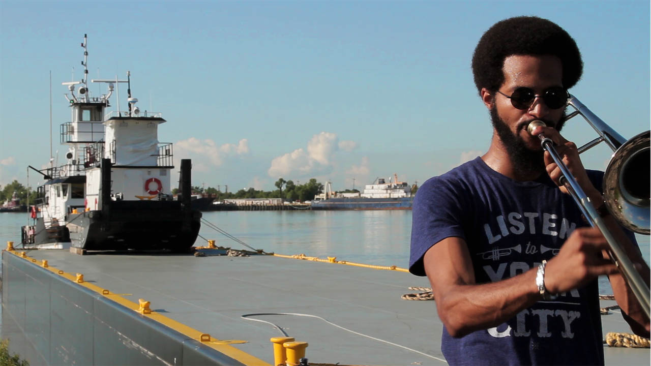 Photograph of a man on pier playing a trombone. A boat is docked in the background.