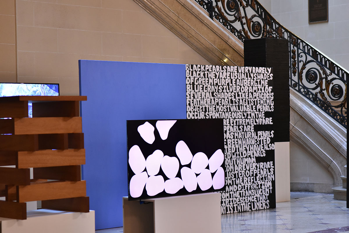 Installation view: Large painting that is one-half half blue color field and one-half white text on a black background. Flat screen monitor and scupture are partially in this view.