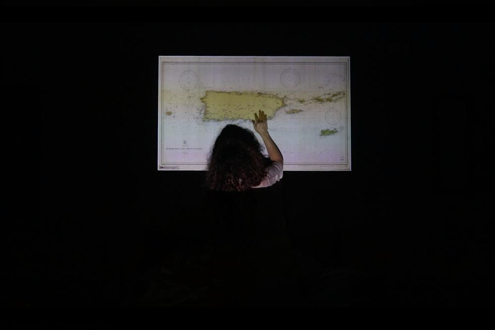 Nude figure in a dark room seen from behind. A map is projected onto the body.
